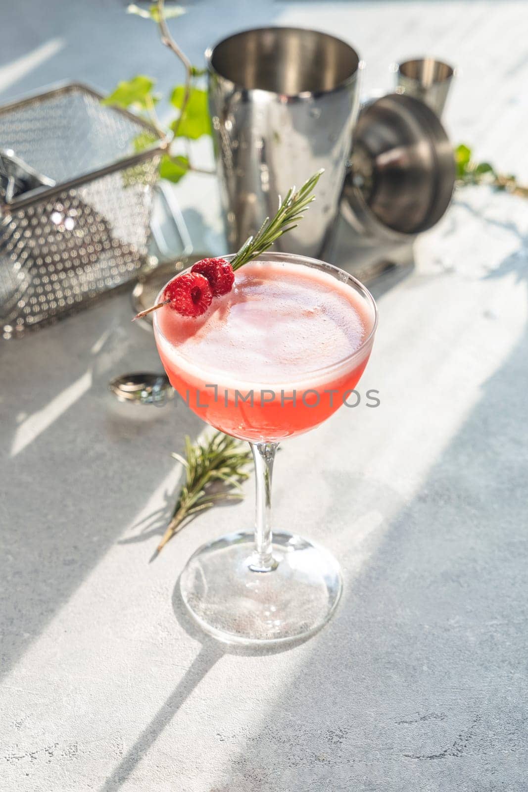 Pink cocktail decorated with raspberries and rosemary sprig, stands on bright table with cocktail shaker and jigger in background