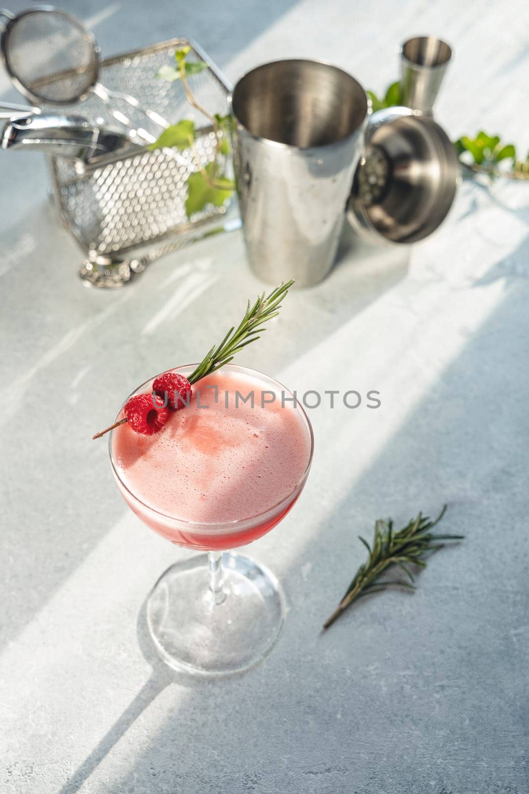 Pink cocktail is garnished with fresh raspberries and rosemary sprig on light gray background with cocktail shaker and jigger out of focus