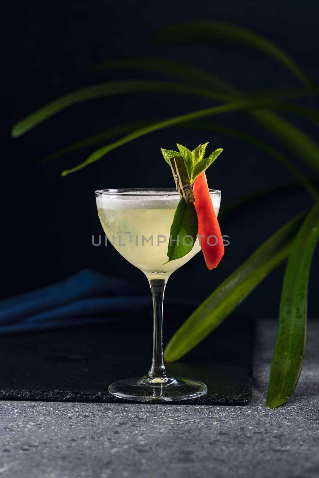Yellow cocktail garnished with a chili pepper and mint leaves, served in a coupe glass on a dark background with tropical plant leaves