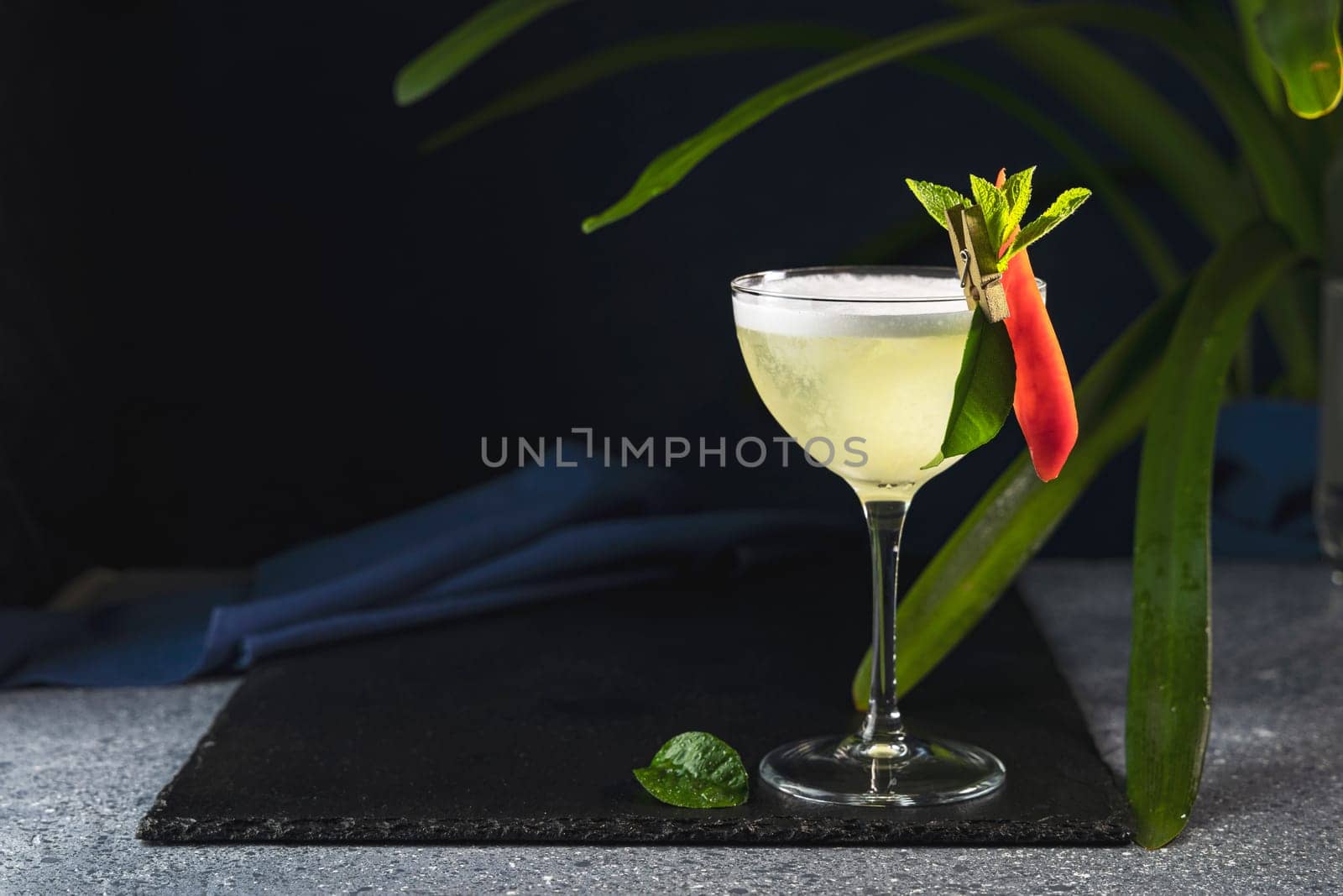 Vibrant yellow cocktail, adorned with a chili pepper and mint, sits on a dark slate, embodying tropical refreshment