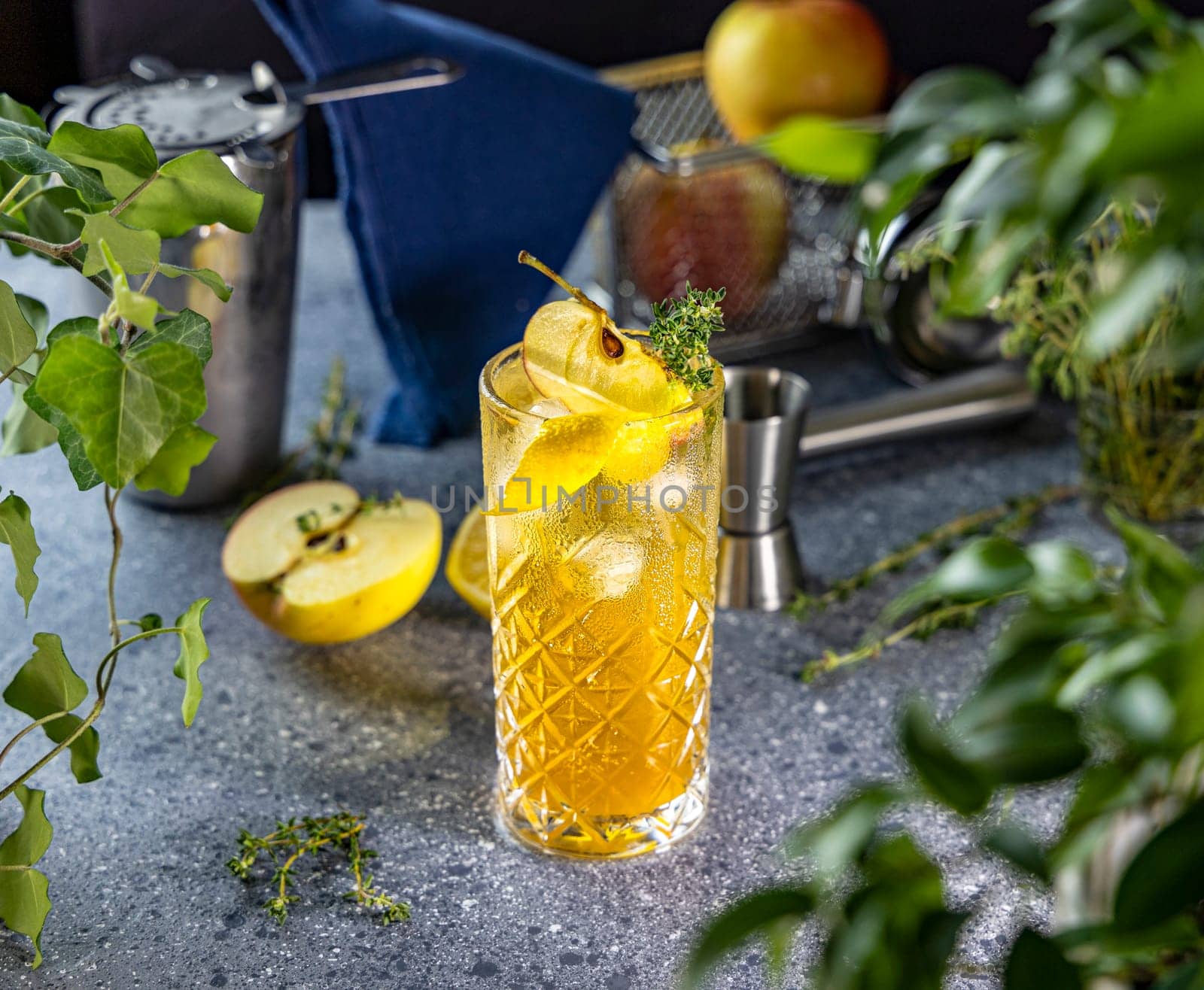 Golden apple cider cocktail in a crystal glass, garnished with fresh apple slices, lemon, thyme sprigs, and ice cubes