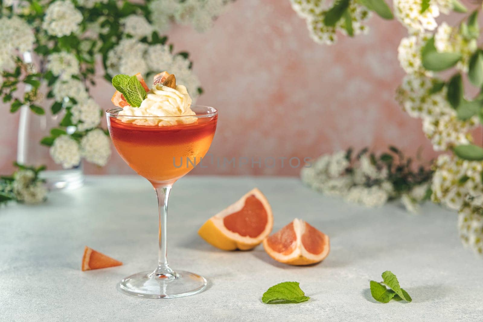 Grapefruit jelly dessert topped with whipped cream and mint, elegantly served in a glass alongside fresh grapefruit slices and flowers on a pink background