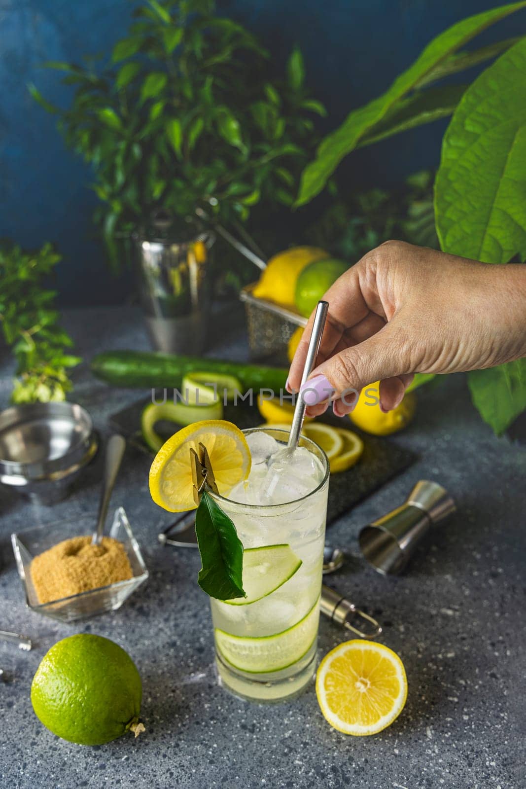 Hand adding metal straw to glass of refreshing homemade lemonade with cucumber, lime, lemon, ice and mint on grey table with ingredients and bar tools