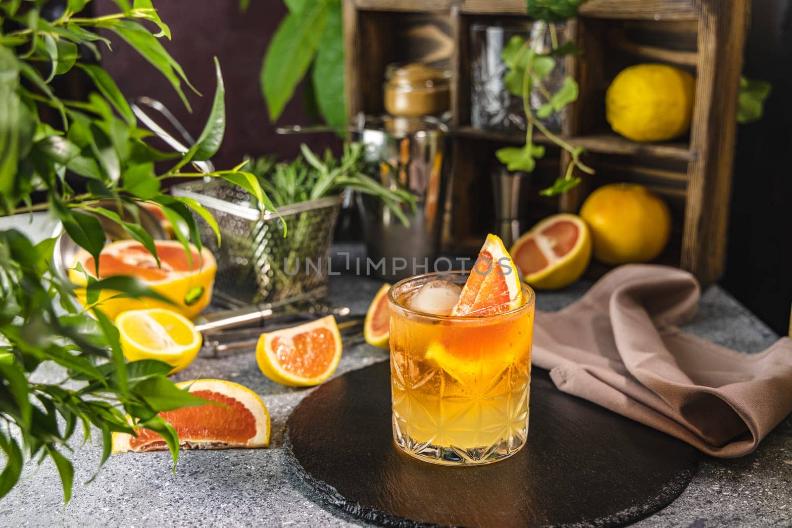 Glass of refreshing orange paloma cocktail is decorated with grapefruit slice and ice cubes, surrounded by fresh citrus fruits and green plants