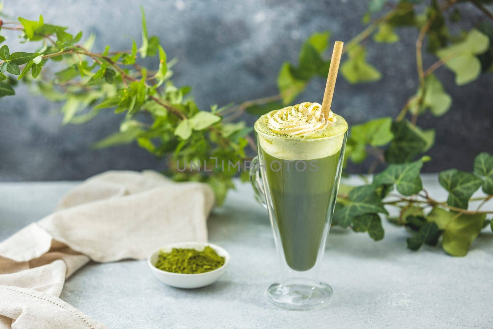 Green tea powder and ivy branches near a tall glass of iced matcha latte with whipped cream and a straw on a table