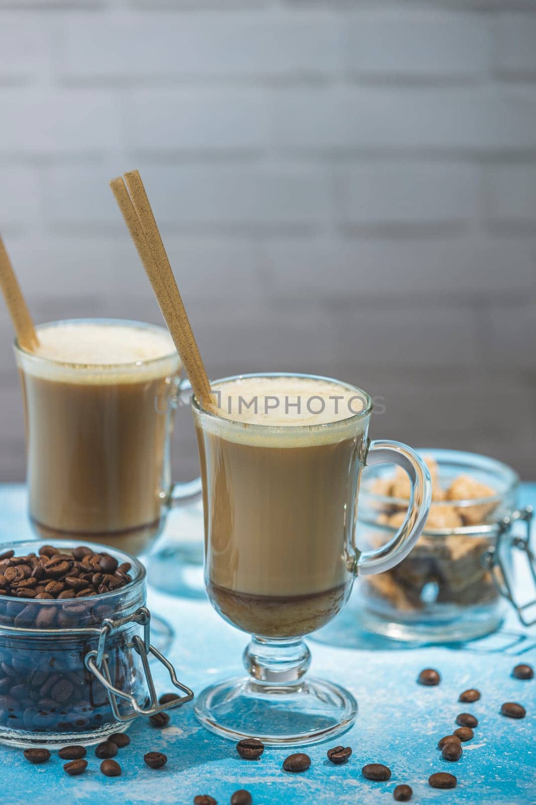Two glasses of latte with eco friendly straws, coffee beans, and brown sugar on a blue table, creating a cozy and inviting coffee break scene