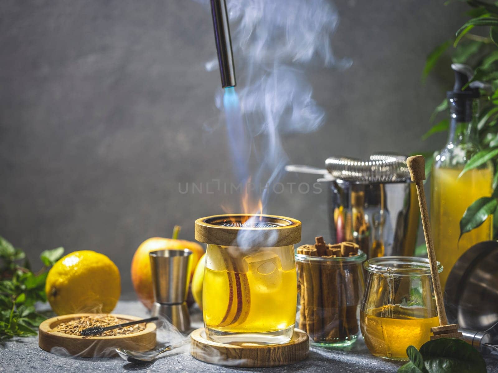 Glass with hot apple cocktail is getting heated by blowtorch in smoky bar setting with ingredients around