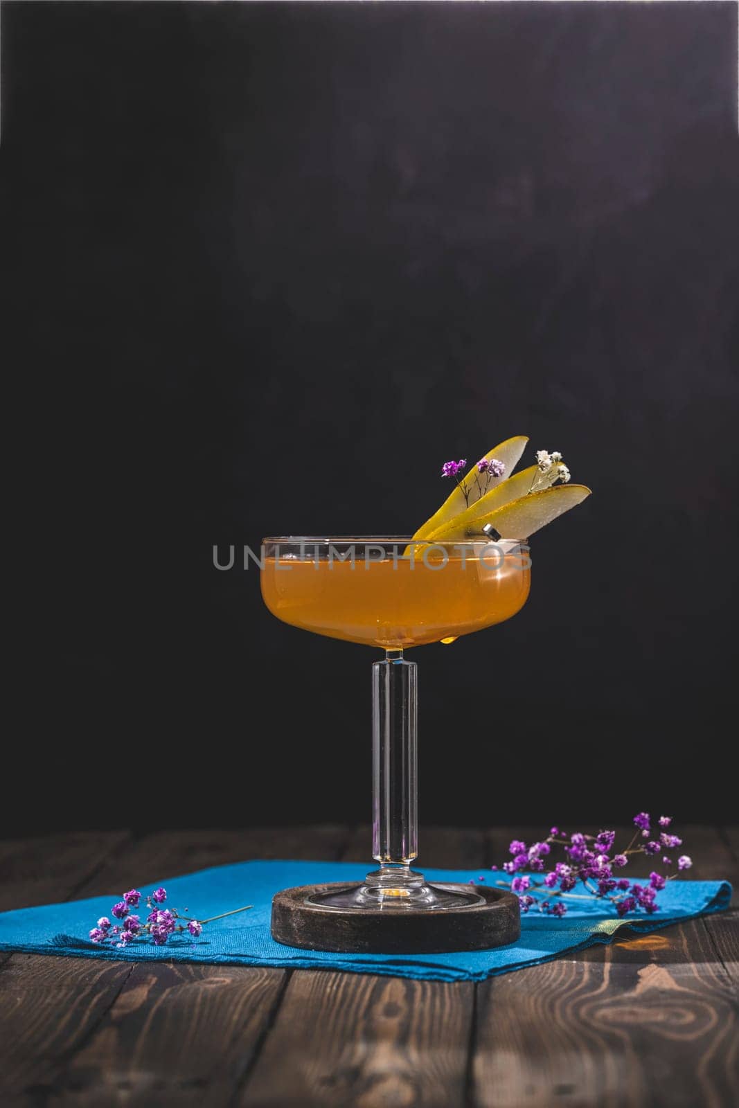 Coupe glass filled with amber liquid, garnished with pear slices and delicate flowers, sits on a dark wooden table with a blue napkin