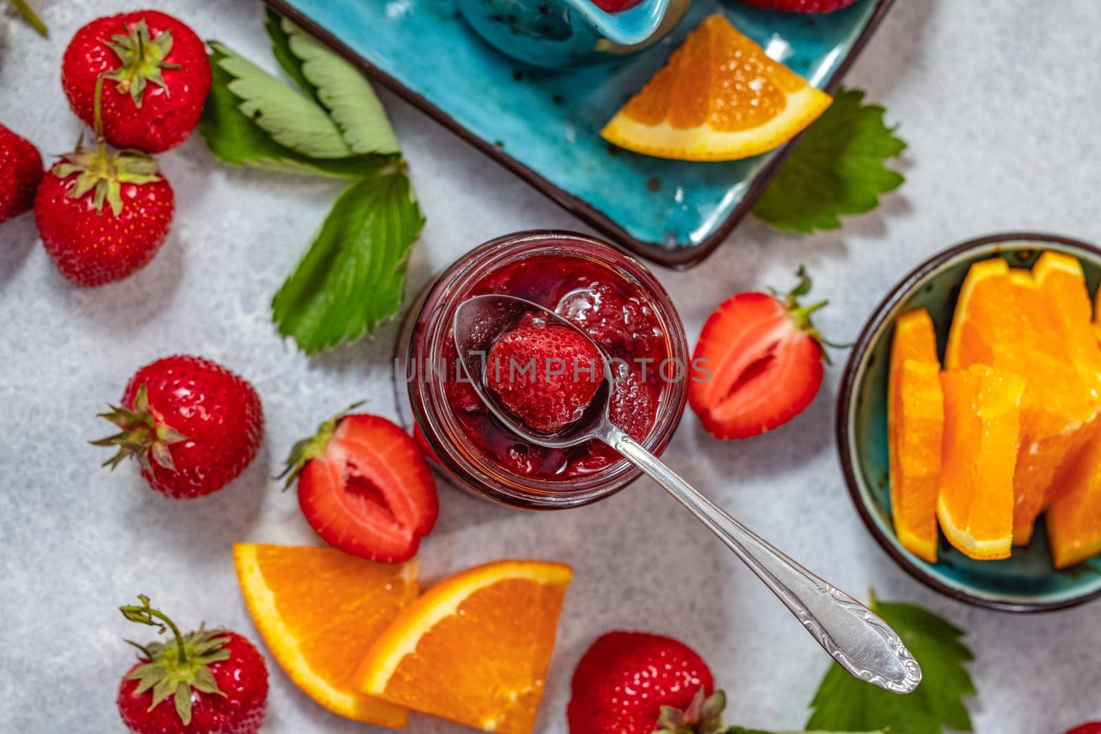 Spoon scooping strawberry from jar of homemade strawberry jam surrounded by fresh strawberries and orange slices