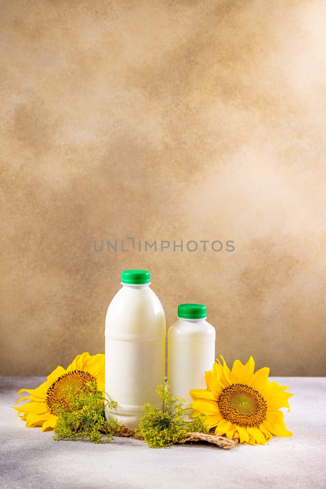 Two white bottles with green caps containing yogurt or milk, decorated with sunflowers, dill, and burlap, rest on a gray table against a brown backdrop