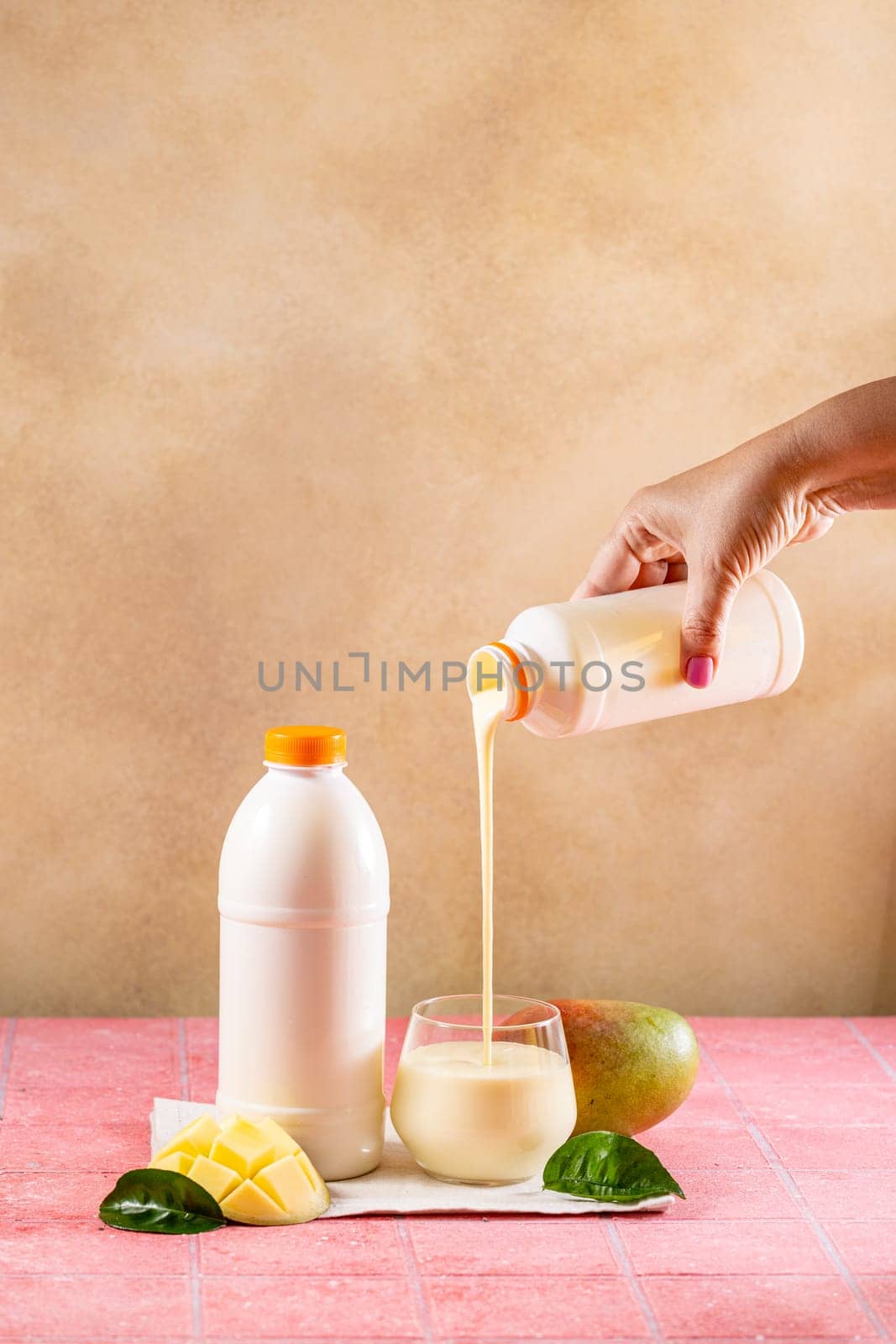 Hand pouring mango lassi or yogurt drink from bottle into glass with fresh mango fruit and leaves on pink tiled table