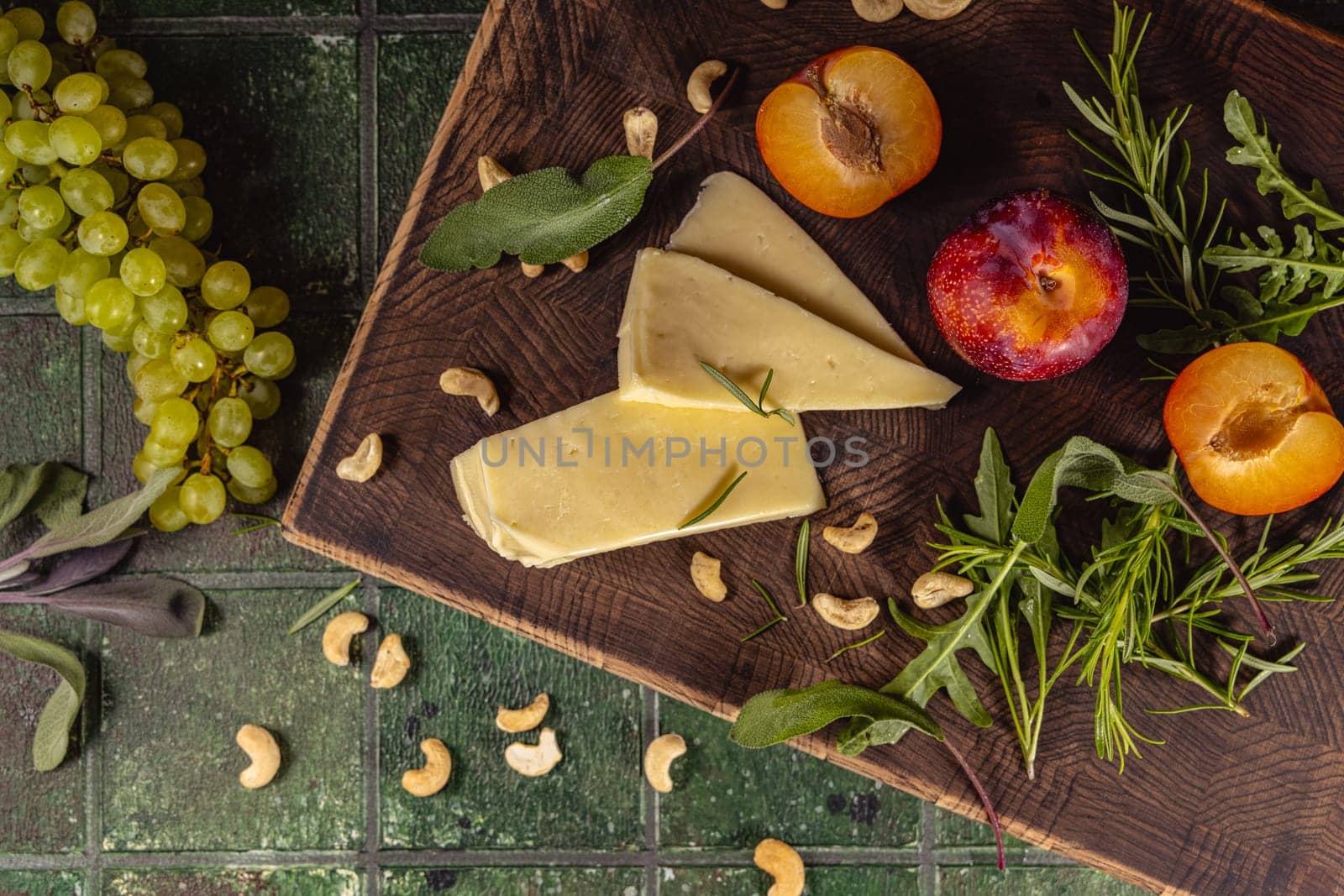 Gourmet cheese selection with fresh grapes, plums, apricots, cashew nuts, and aromatic herbs, creating an enticing culinary scene on a rustic wooden board