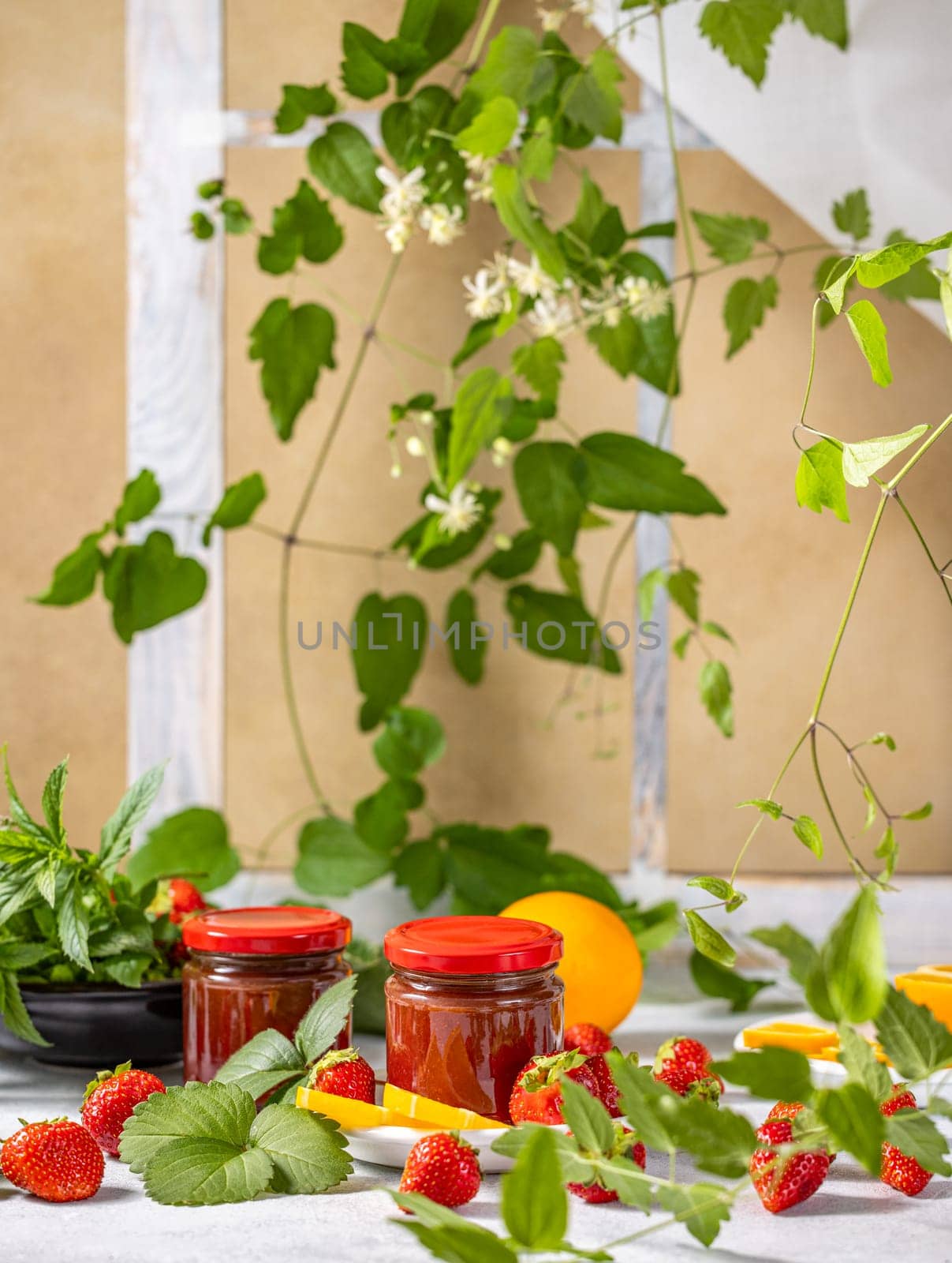 Homemade strawberry jam in glass jars with fresh strawberries, orange slices, mint leaves, and climbing plant on a table