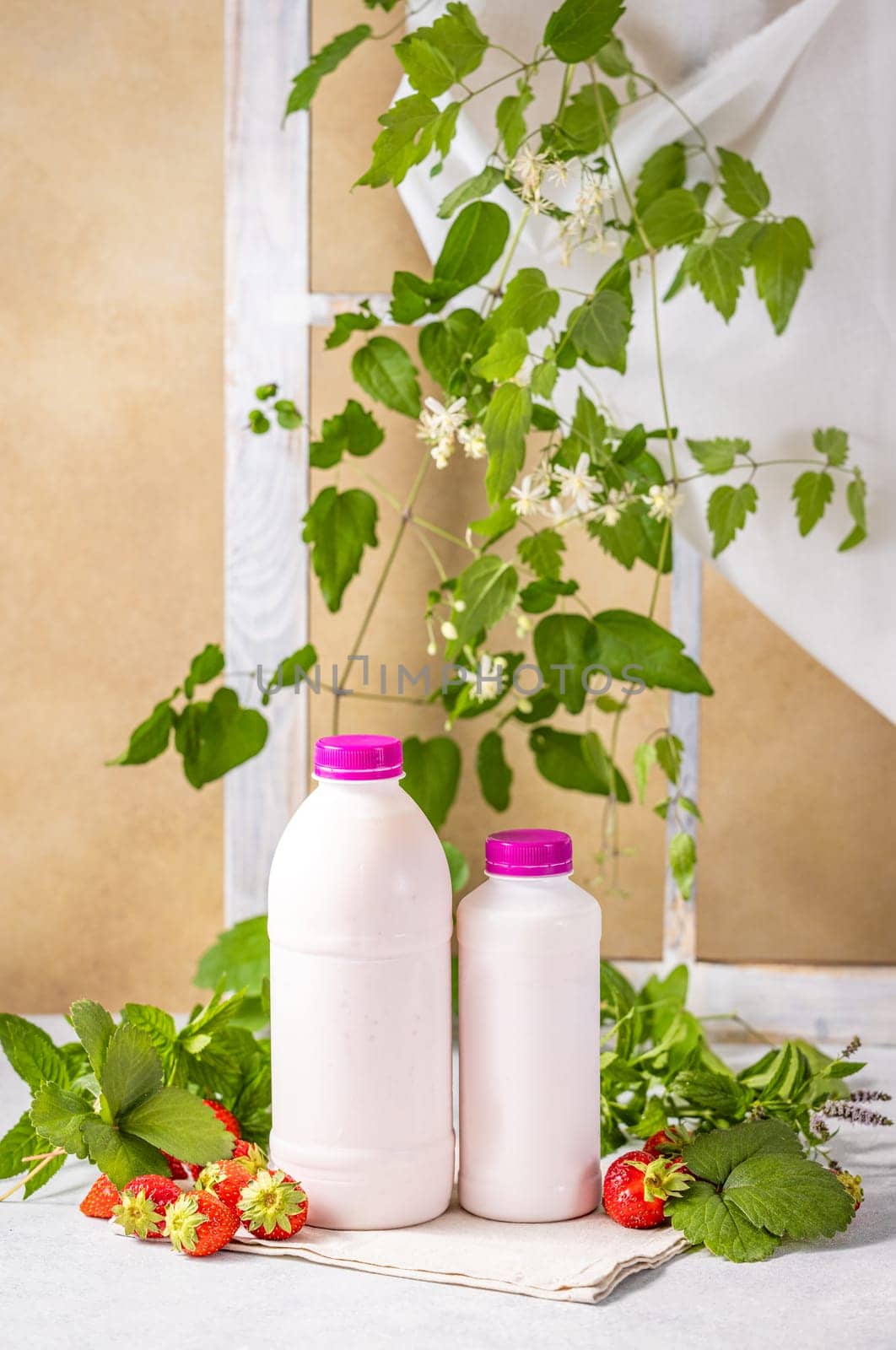 Two plastic bottles of strawberry yogurt are placed on a table with fresh strawberries, mint leaves, and plants, creating a refreshing and healthy scene