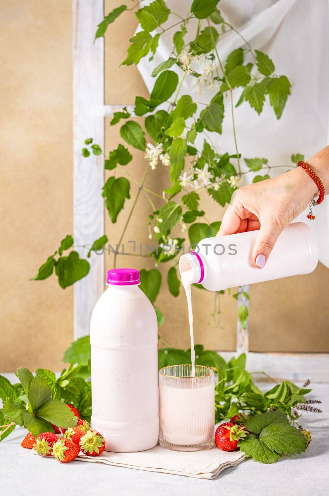 Hand pouring strawberry yogurt from bottle into glass, with fresh strawberries, mint, and decorative plants, promoting healthy eating and refreshment