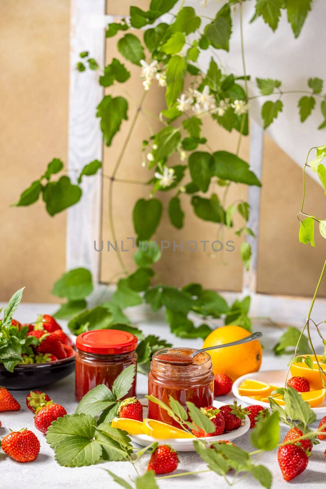 Jars of strawberry jam surrounded by fresh strawberries, orange slices, and green leaves create a vibrant and appetizing scene