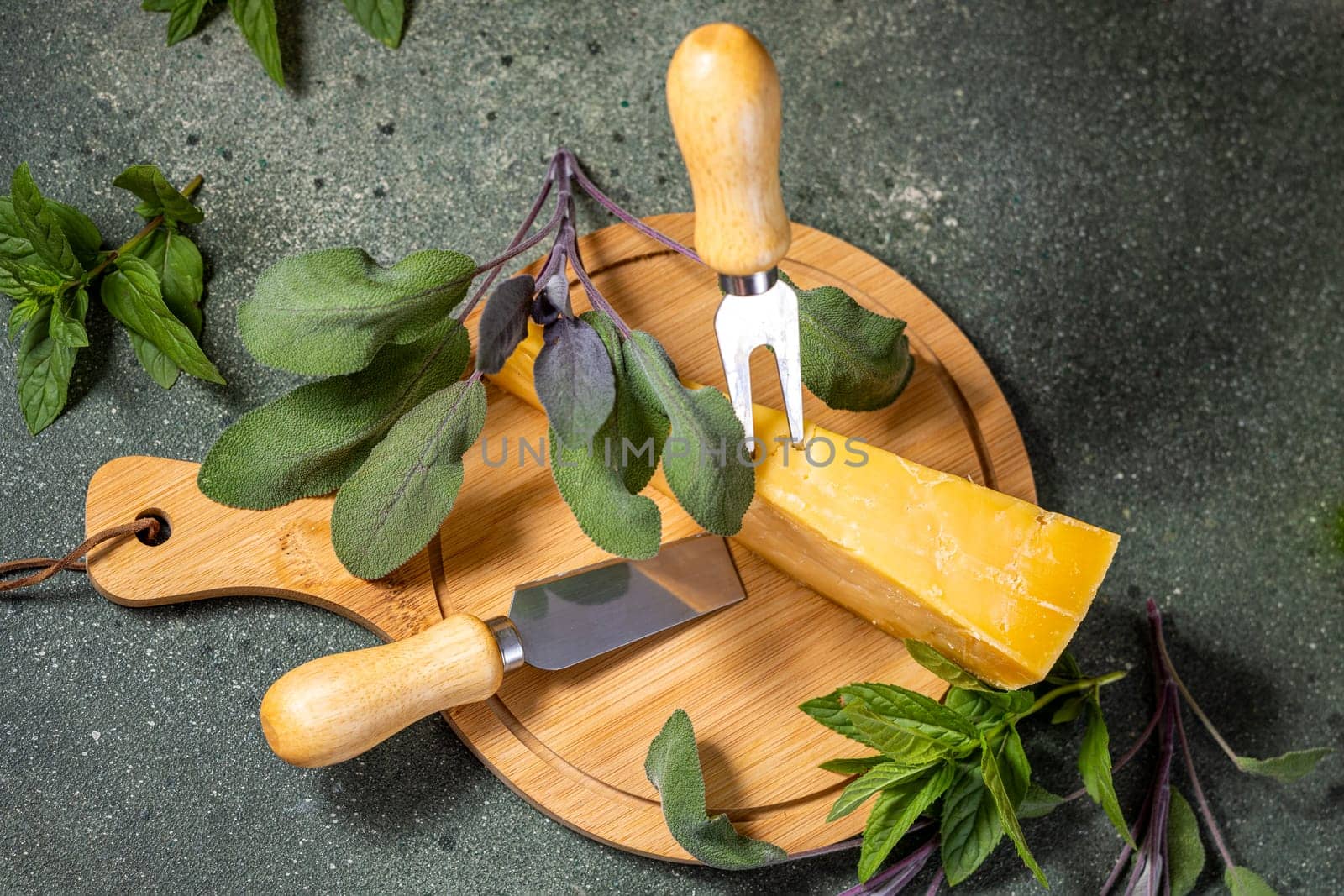 Delicious hard cheese served on round wooden cutting board with cheese knife, fork and fresh mint and sage