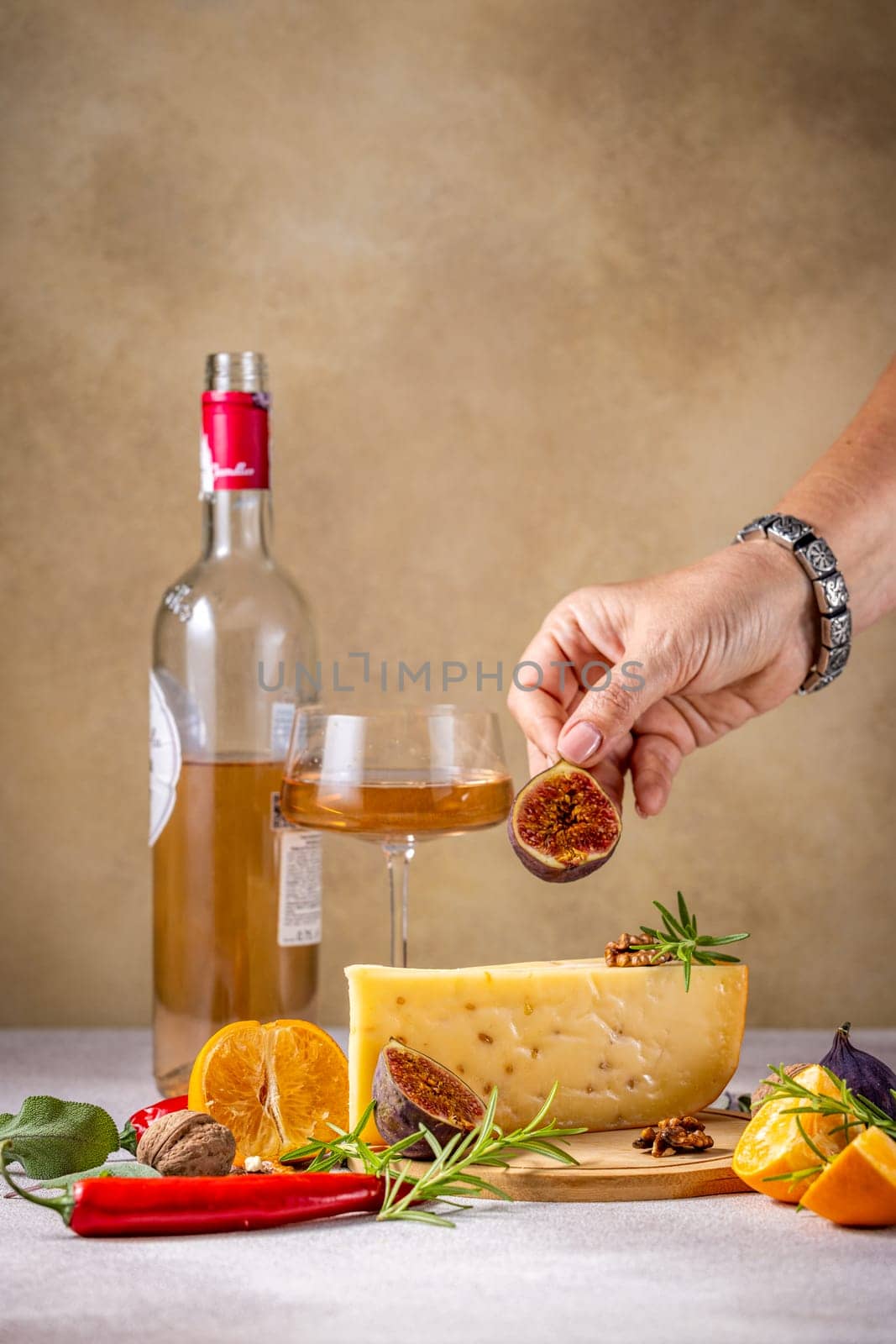 Hand adding a fig slice to a cheese wedge, accompanied by a glass of wine, fruits, and spices, creating a gourmet food and drink setting