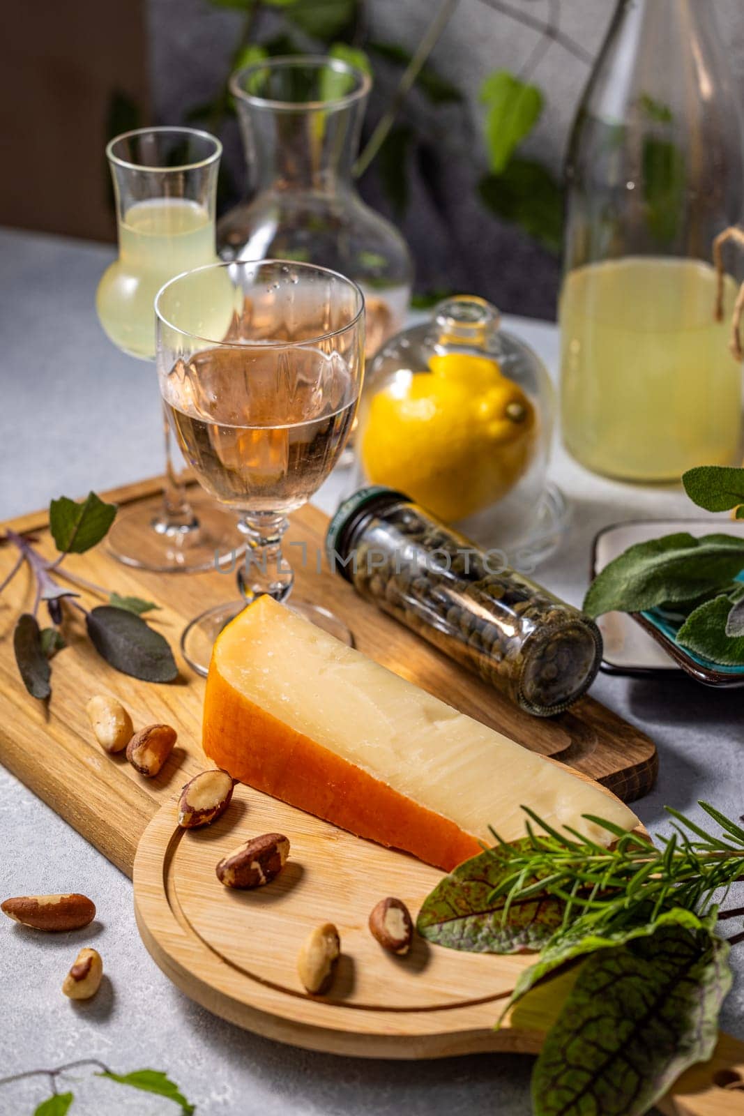 Wedge of cheese, accompanied by brazil nuts, rests on a wooden board, complemented by glasses of rose wine, limoncello, and various herbs and spices, creating a sophisticated culinary arrangement