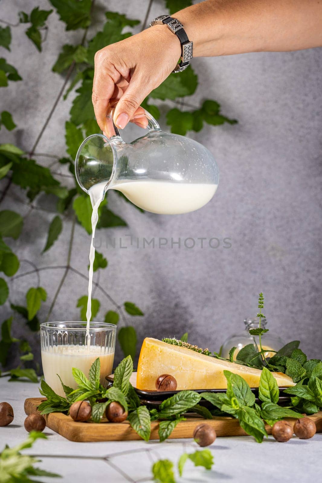 Woman pouring milk from glass jug into a glass, with aged cheese, macadamia nuts and mint leaves on wooden board