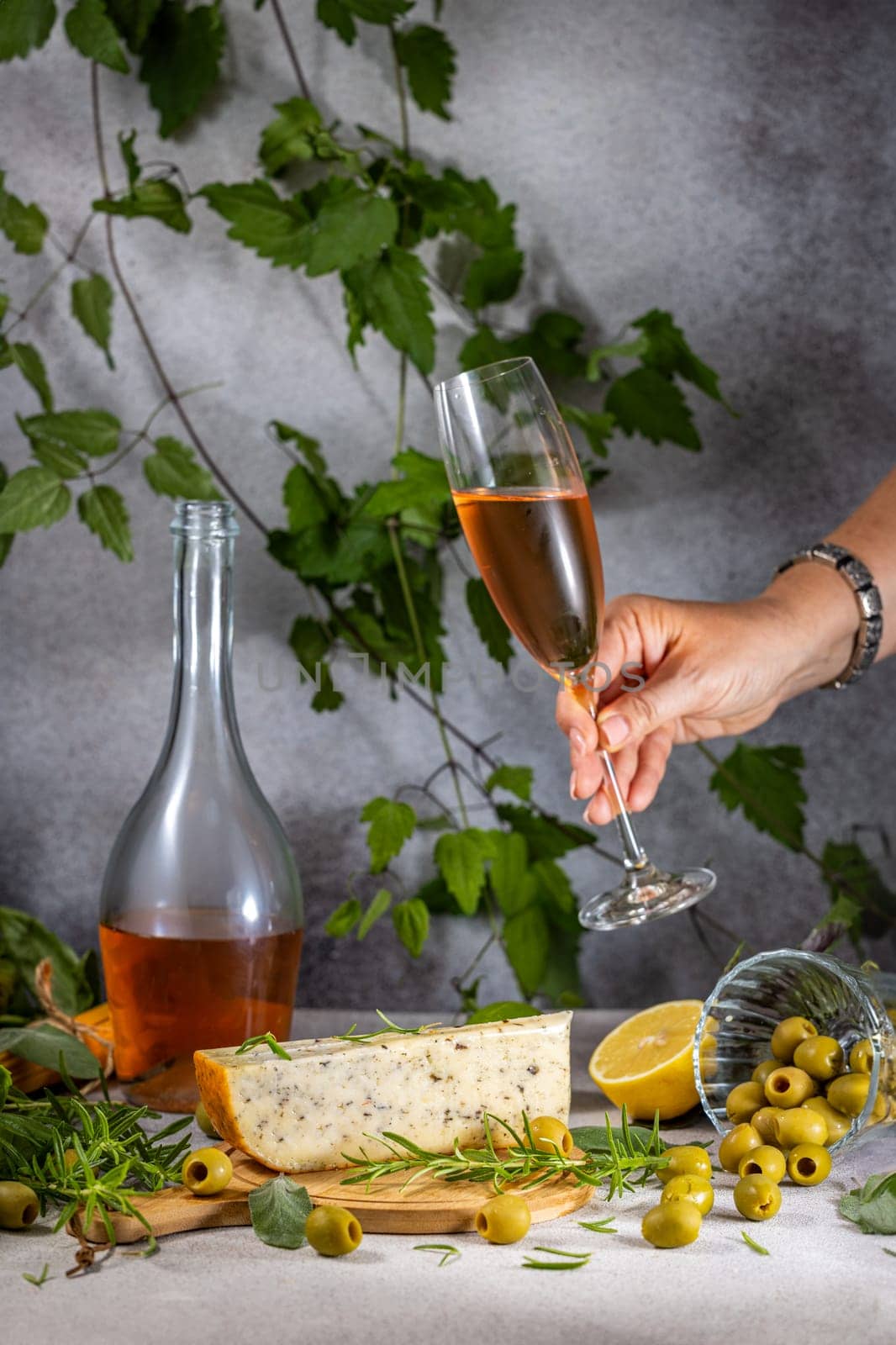 Hand holding a glass of rose wine with gourmet cheese, olives, lemon, and rosemary garnish, creating an elegant appetizer pairing