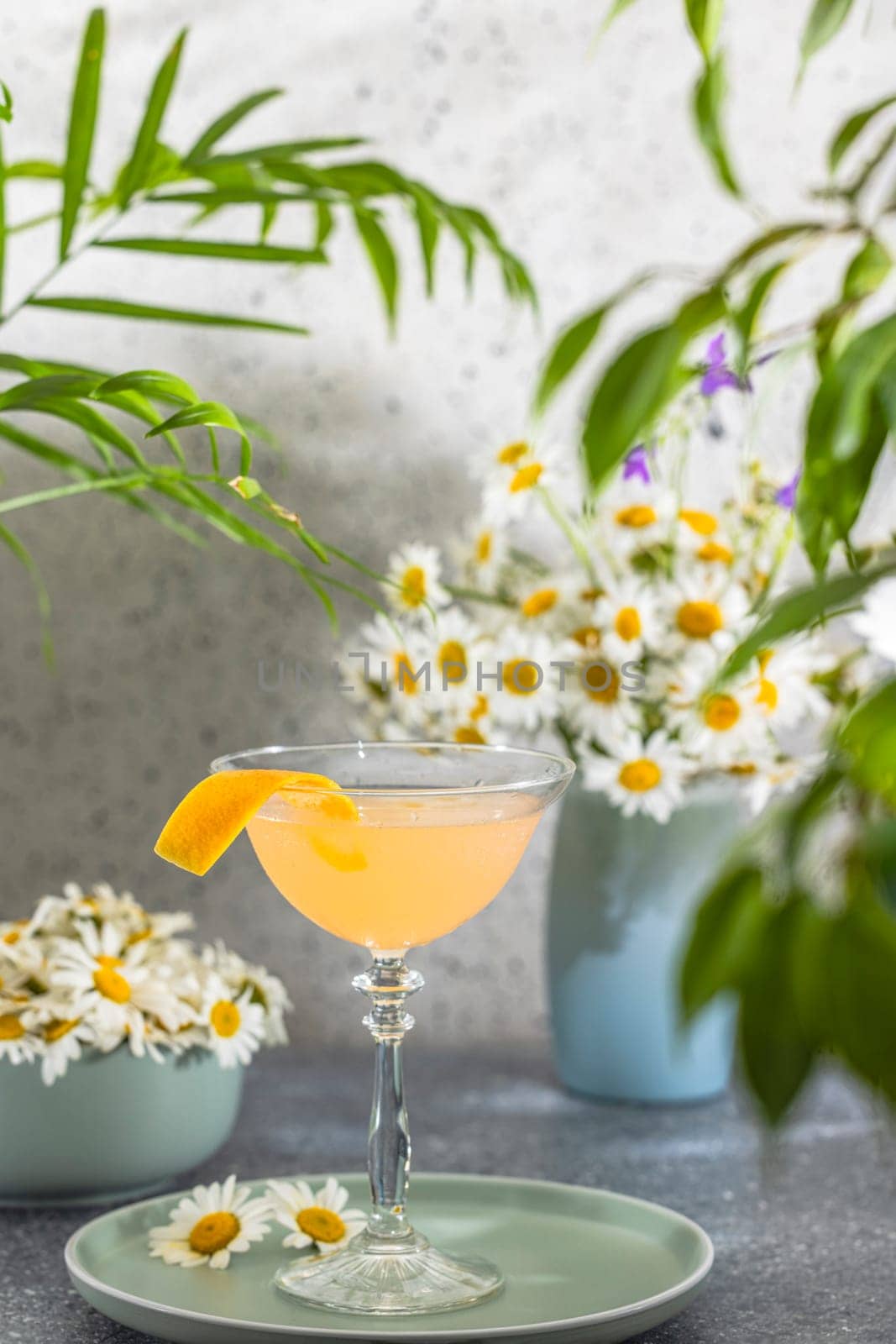 Orange cocktail sits on a table, garnished with an orange peel, surrounded by daisies and lush greenery, creating a refreshing summer scene