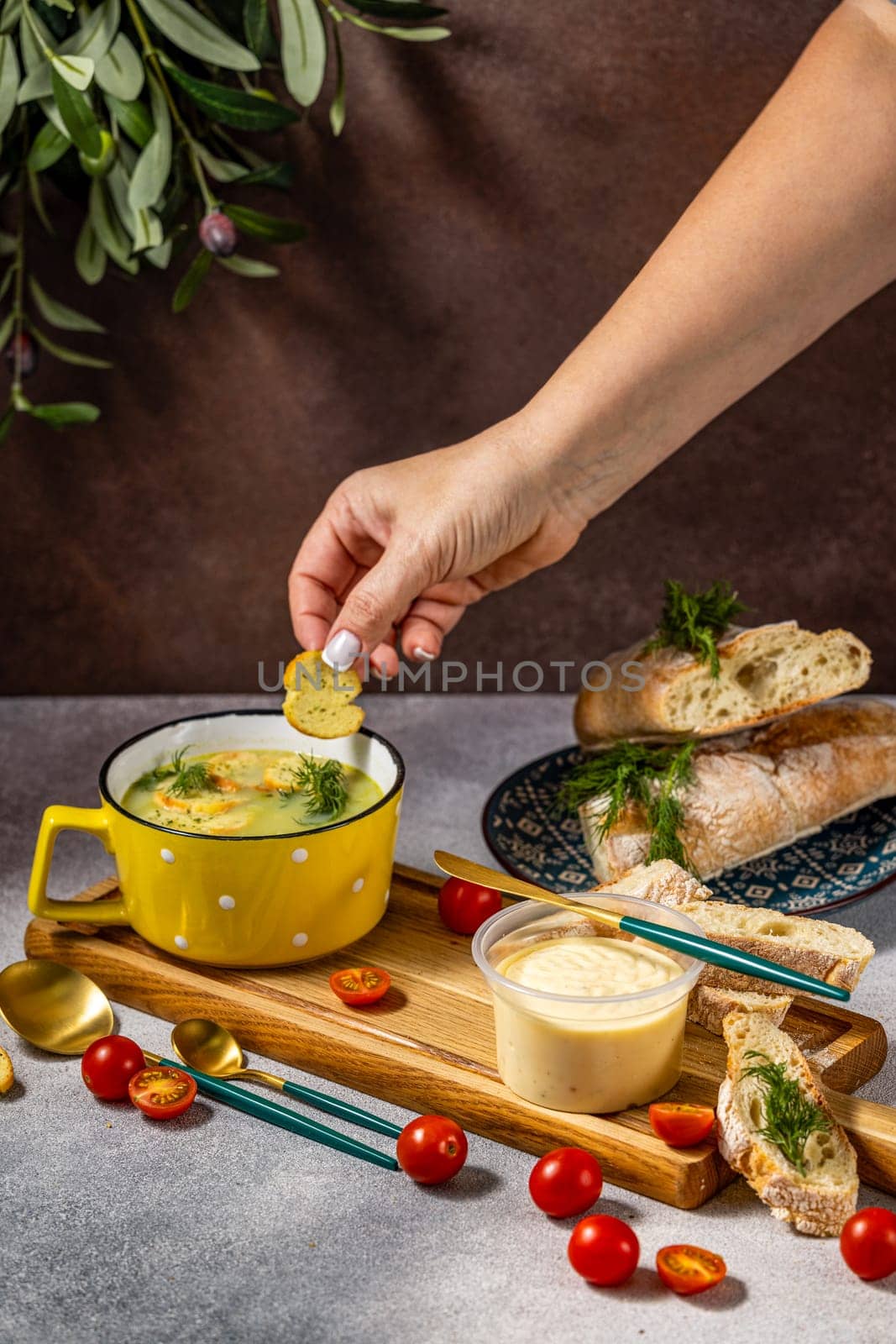 Hand adding croutons to a creamy green pea soup, served with toasted slices of baguette, cherry tomatoes, and a side of sauce
