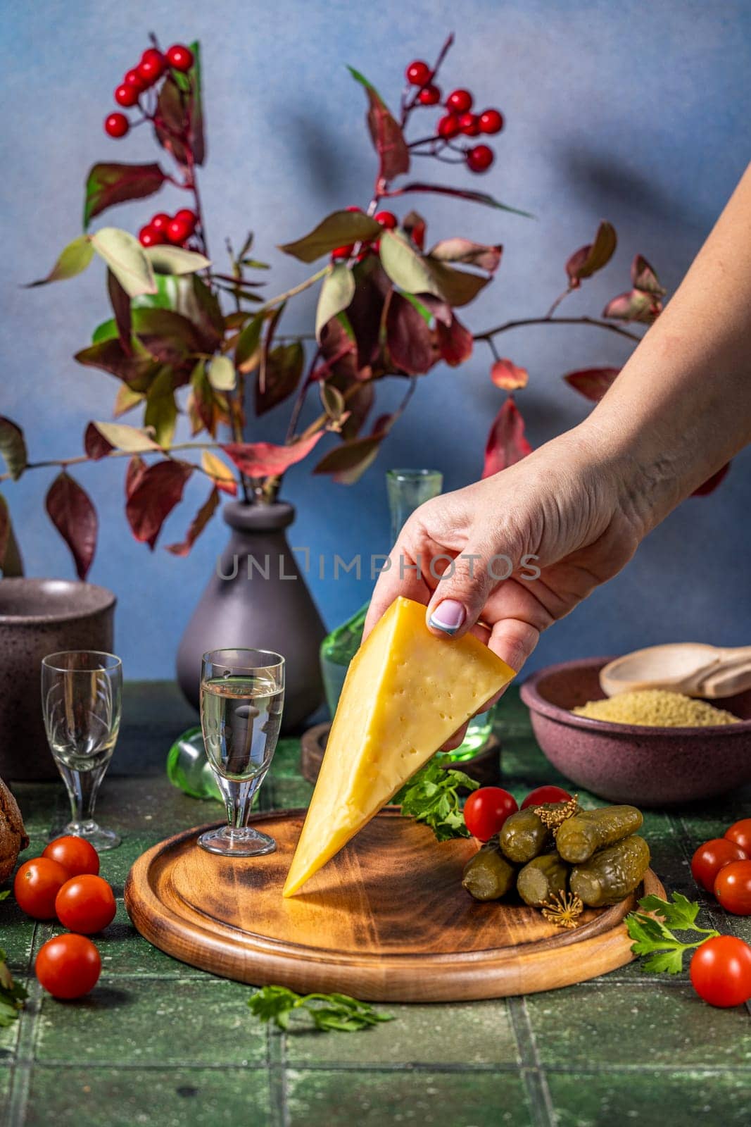 Hand placing cheese on a wooden board with traditional russian snacks like pickles, cherry tomatoes, and vodka shots