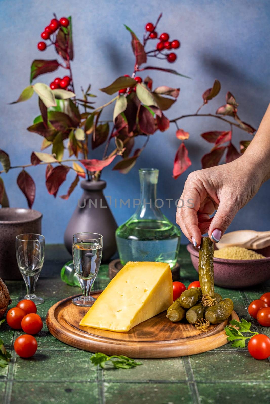 Hand placing a gherkin on a wooden board with cheese, accompanied by vodka shots, cherry tomatoes, and couscous, creating a rustic still life