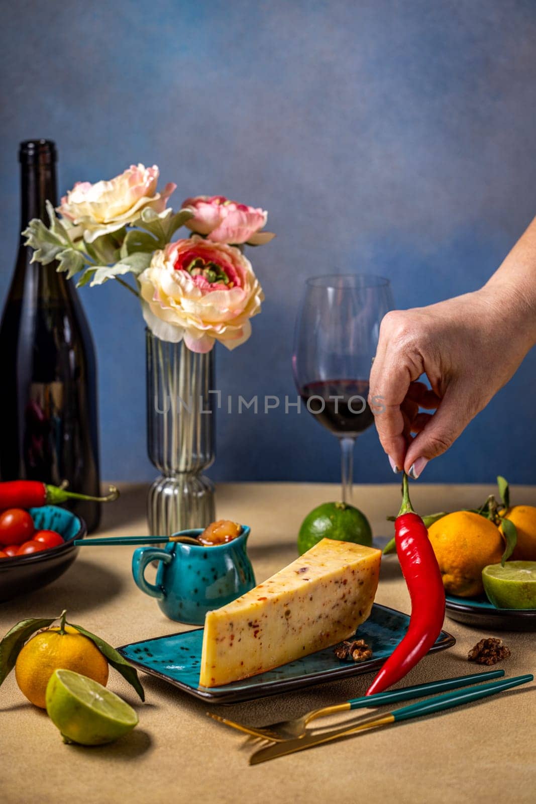 Hand placing a chili pepper on a cheese board, alongside a glass of red wine, various fruits, and a small bowl of jam