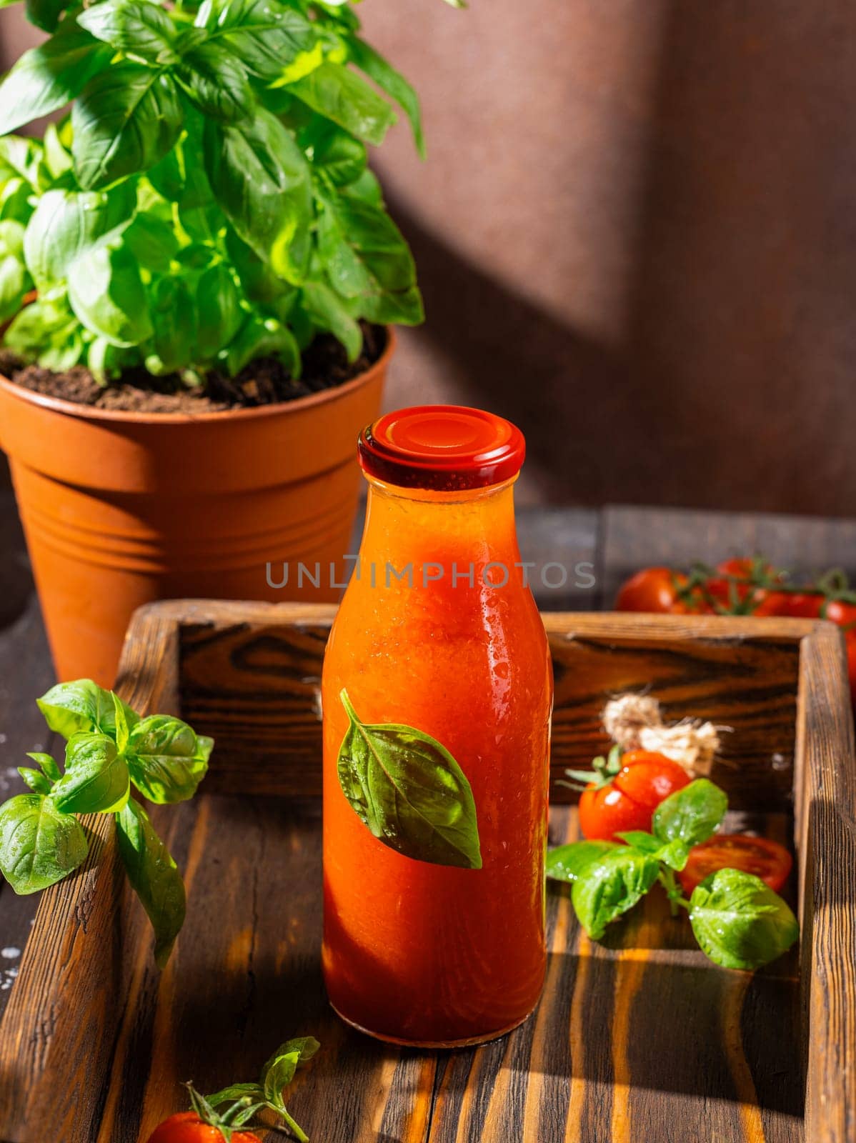 Glass bottle of refreshing tomato juice with basil leaves, accompanied by fresh tomatoes and a basil plant, promoting healthy and natural beverages