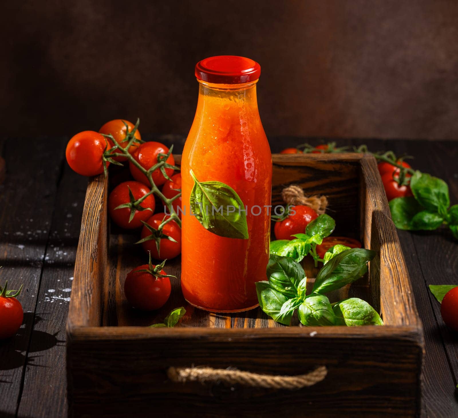 Glass bottle of refreshing tomato juice with basil leaves and ripe cherry tomatoes on a rustic wooden tray, creating a vibrant and healthy image
