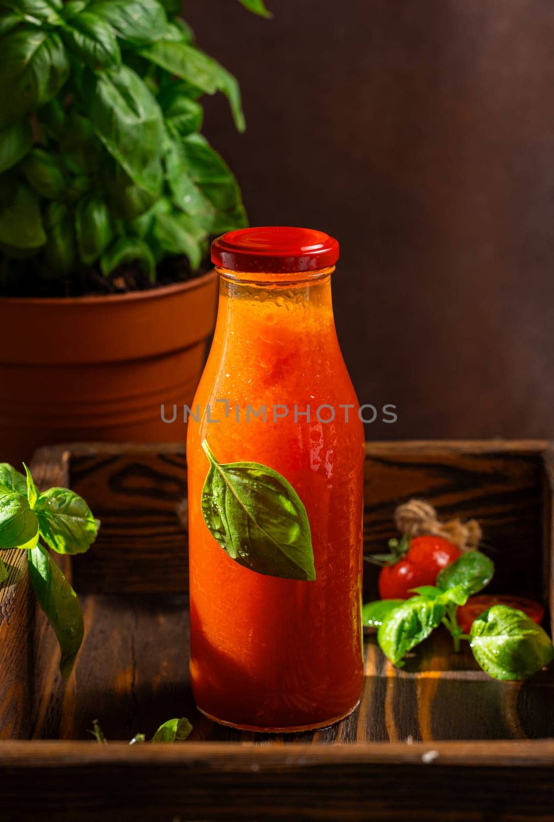 Glass bottle of refreshing tomato juice decorated with basil leaf, placed on wooden tray, promoting healthy and natural beverage options