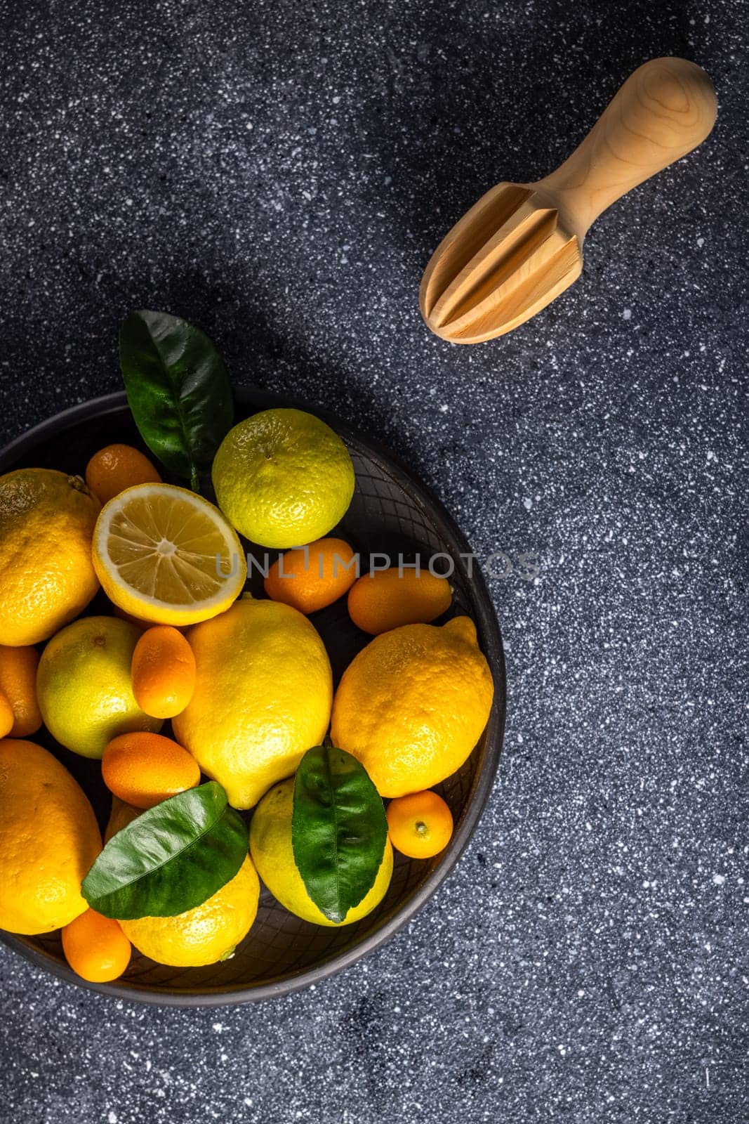 Vibrant citrus fruits like lemons, limes, and kumquats arranged in a black bowl alongside a wooden reamer, creating a refreshing scene on a dark stone tabletop