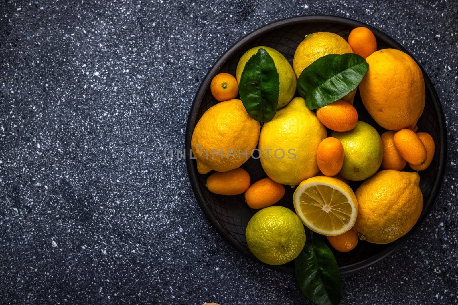 Vibrant citrus fruits, including lemons, limes, kumquats, and oranges, create a colorful arrangement on a dark plate, showcasing their freshness and variety