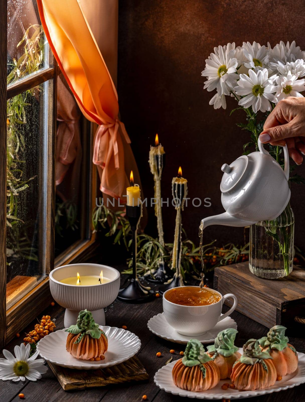 Cozy autumn scene with pumpkin shaped desserts, a woman pouring tea, and candles by a window on a rainy day