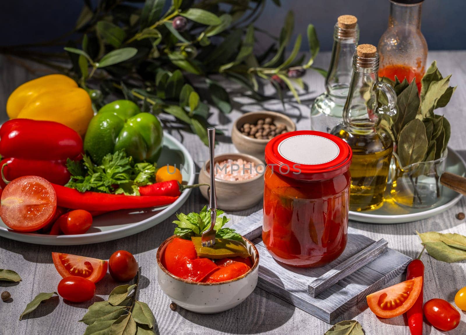 Glass jar of homemade preserved tomatoes, surrounded by fresh tomatoes, peppers, spices, herbs, and olive oil, showcasing the ingredients and the final product