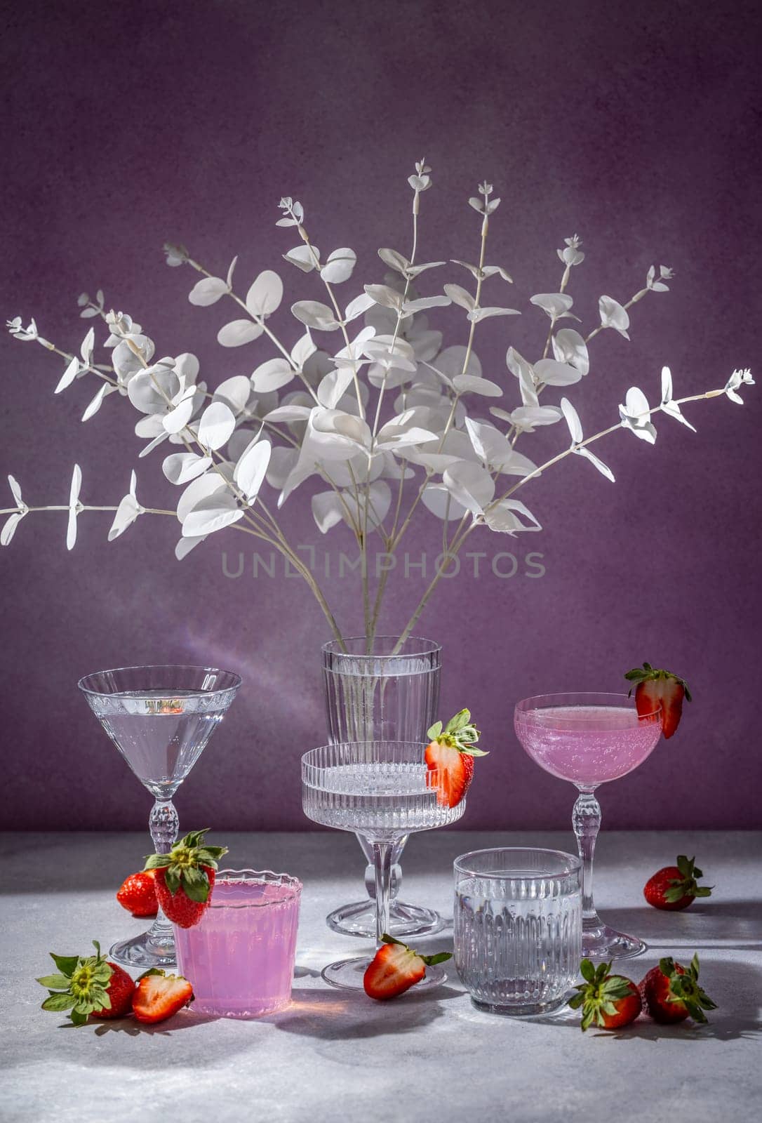 Pink cocktails and elegant glassware are beautifully arranged with fresh strawberries and white eucalyptus branches, creating a stylish and refreshing scene