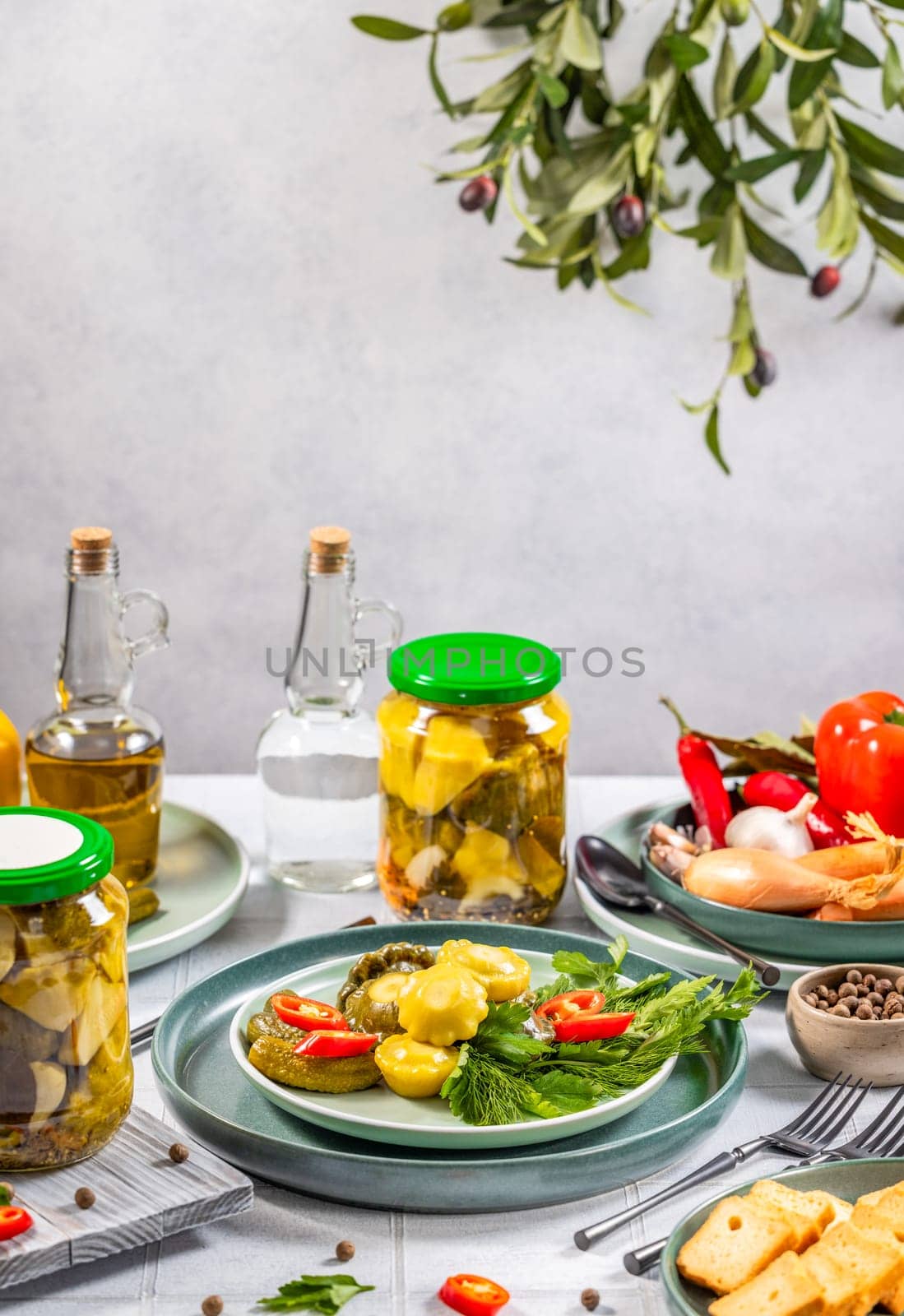 Pickled pattypan squash and gherkins on a plate, garnished with fresh herbs, alongside jars of pickles, oil, and spices