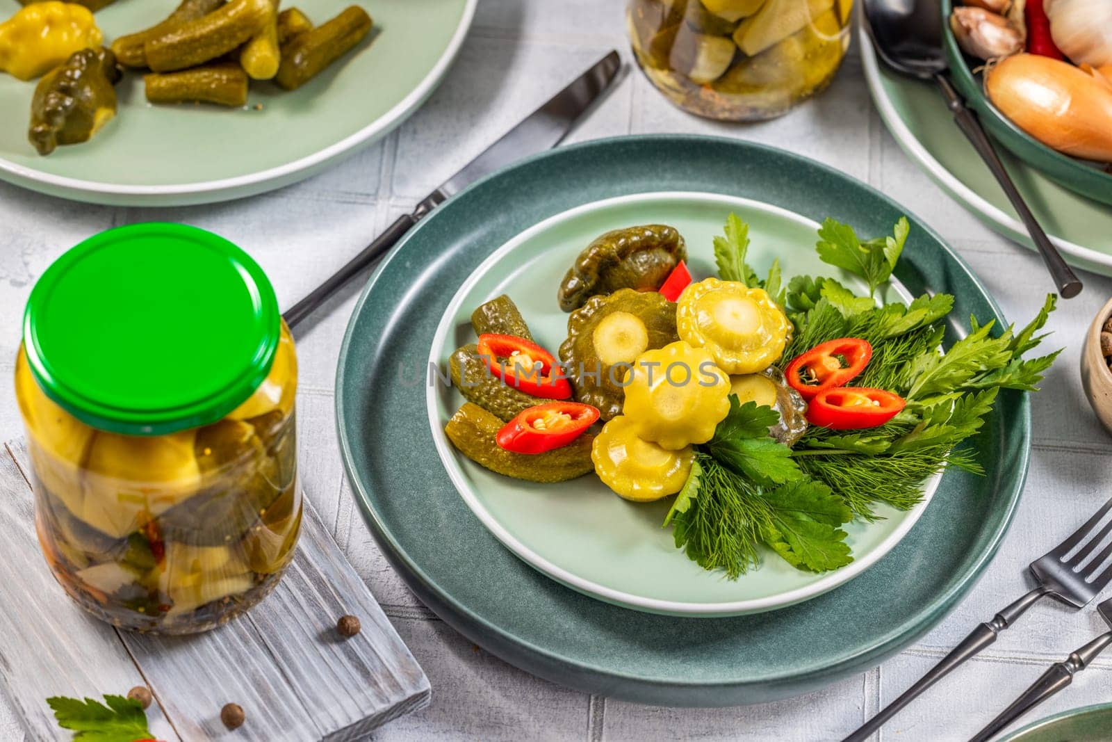 Pickled vegetables, pattypan squash, cucumbers, and chili peppers, served on a green plate with fresh herbs, dill, and parsley