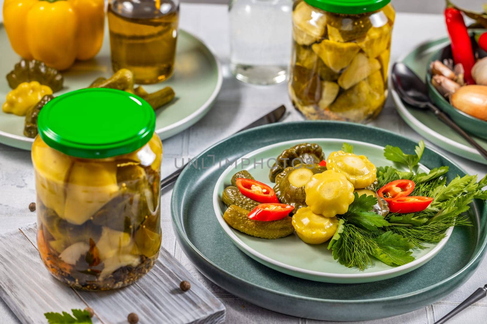 Pickled pattypan squash, gherkins, chili pepper and herbs served on a plate, with jars of pickles and other ingredients in the background