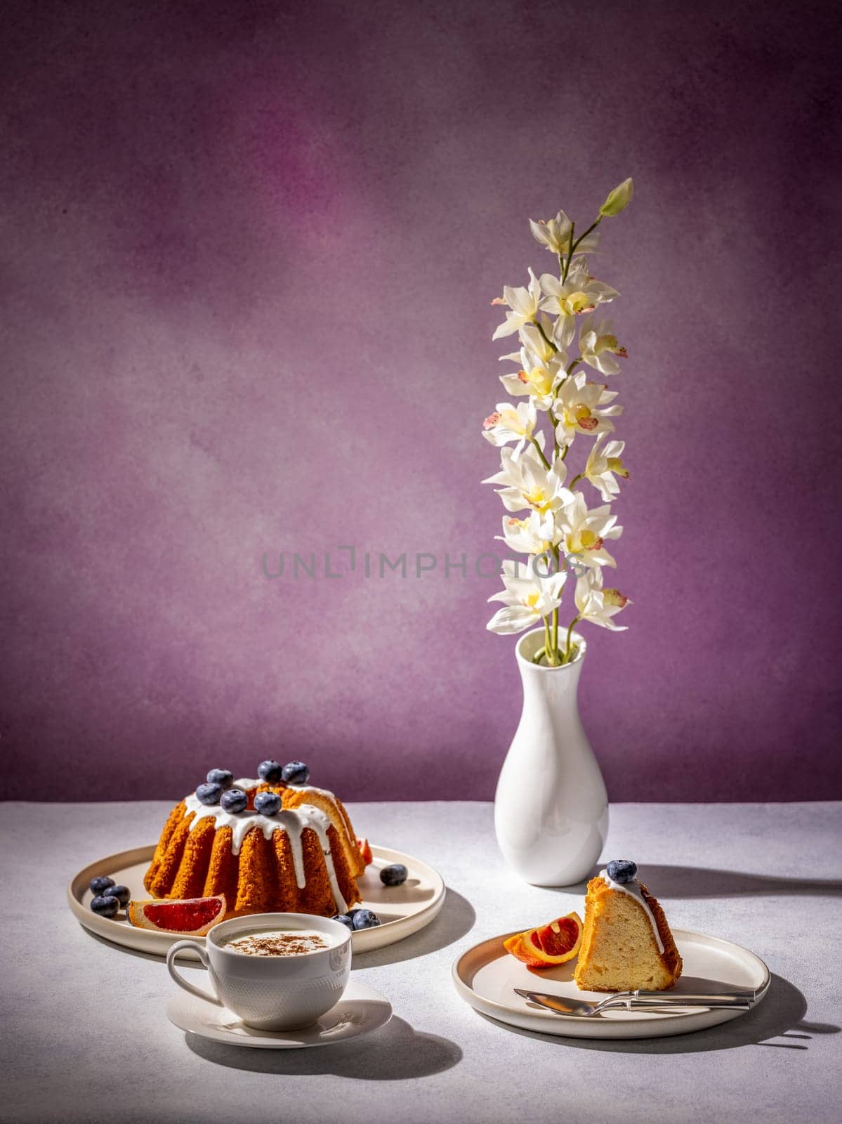 Gugelhupf cake with icing, blueberries, orange slices, cappuccino and orchid flower in a vase, creating a cozy and inviting atmosphere
