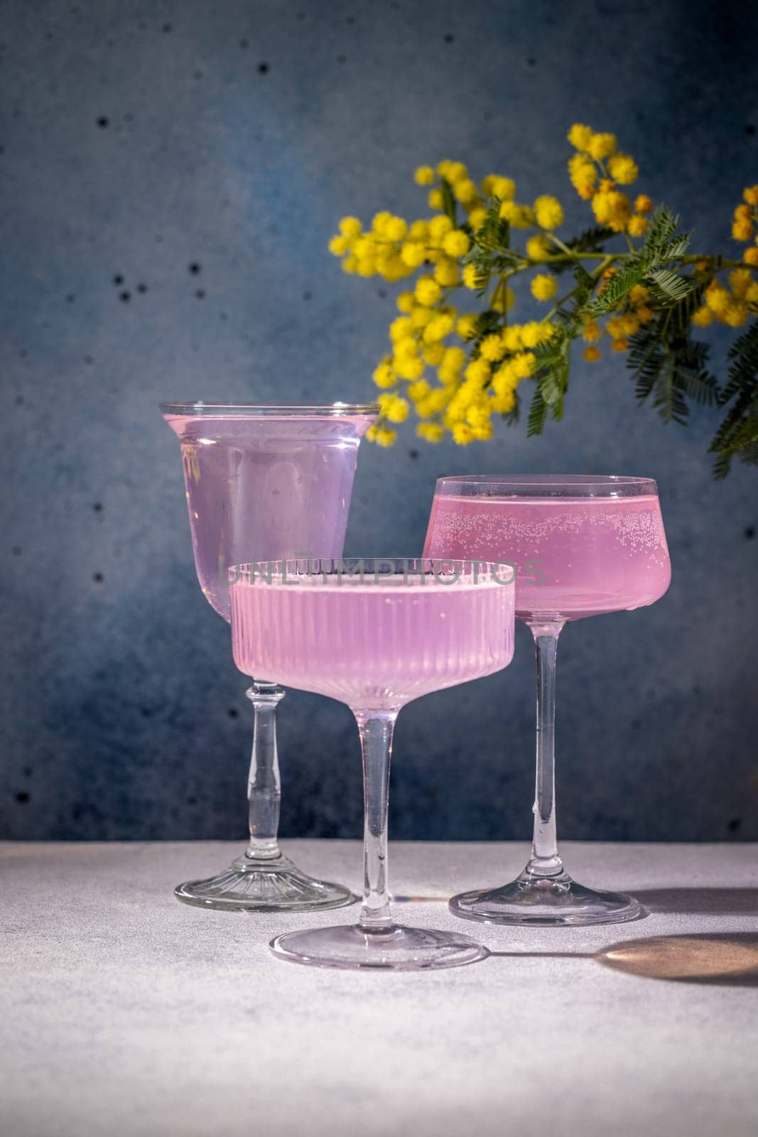 Three glasses of pink cocktails stand on a table with mimosa flowers in the background, creating a stylish and festive atmosphere