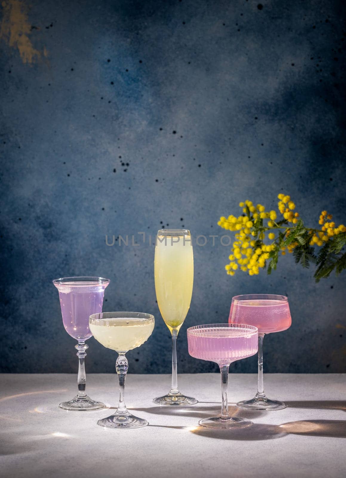 Four elegant glasses filled with vibrant cocktails, accompanied by a mimosa flower, create a celebratory atmosphere, perfect for special occasions