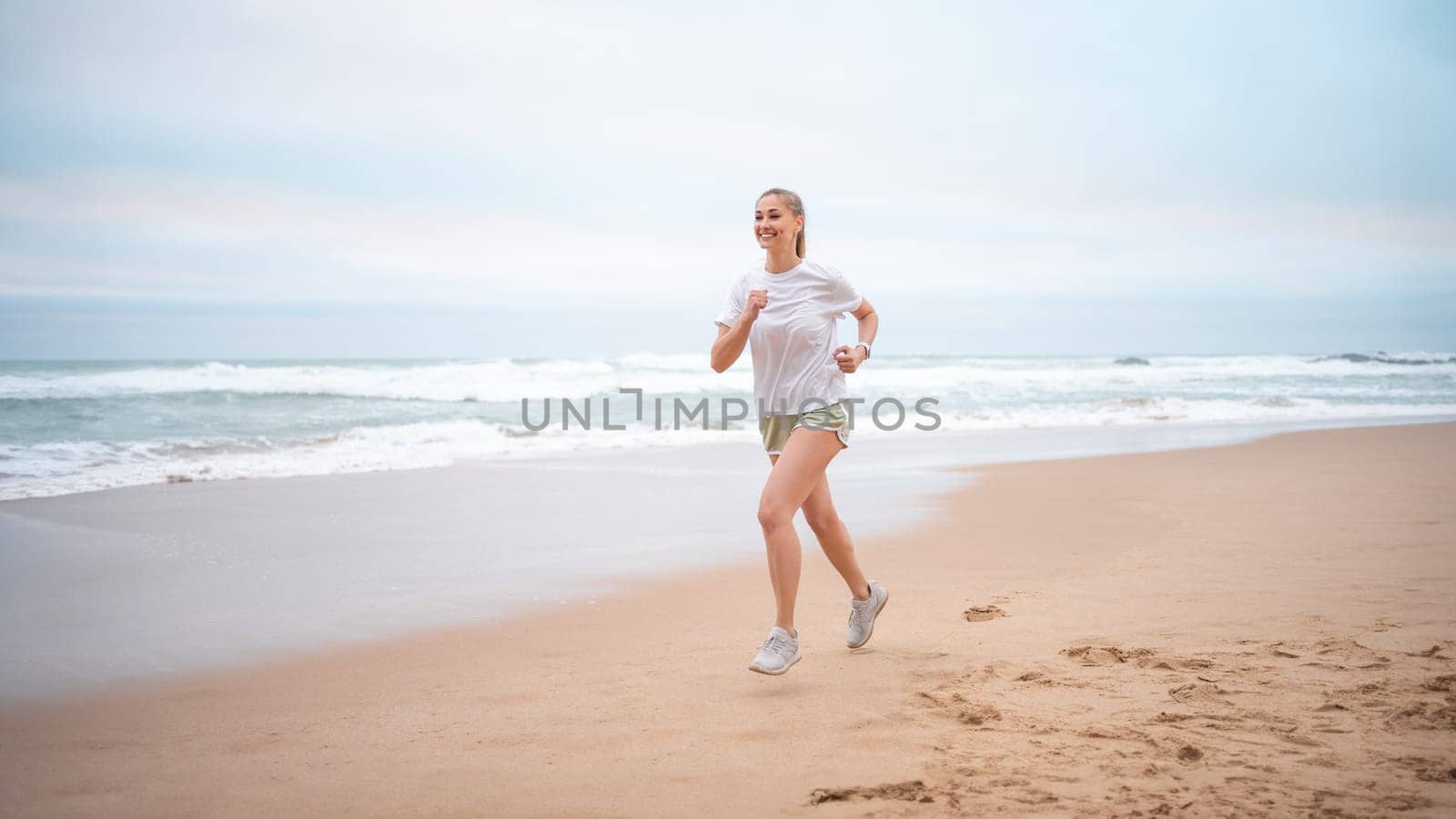 Happy sporty woman jogging on wet seashore against sky at beach. Full body of smiling female in sportswear exercising during daytime