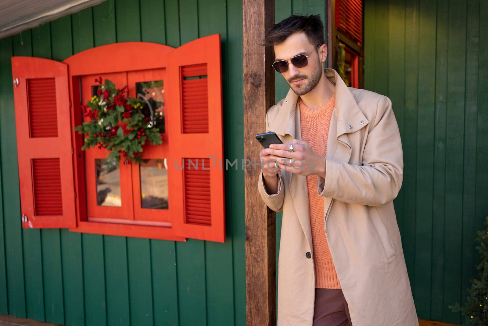 Handsome man in beige trench coat using smartphone outside green house. Young male is leaning on wooden pole during daytime