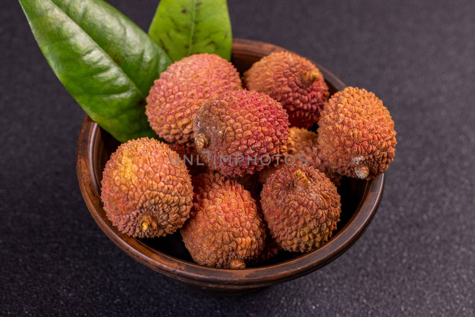 Ripe lychees and green leaves presented in a wooden bowl, creating a vibrant display against a dark backdrop