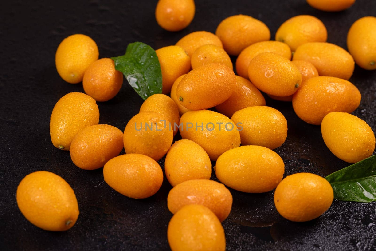 Vibrant kumquats, adorned with water droplets and accompanied by green leaves, create a visually appealing display on a dark surface