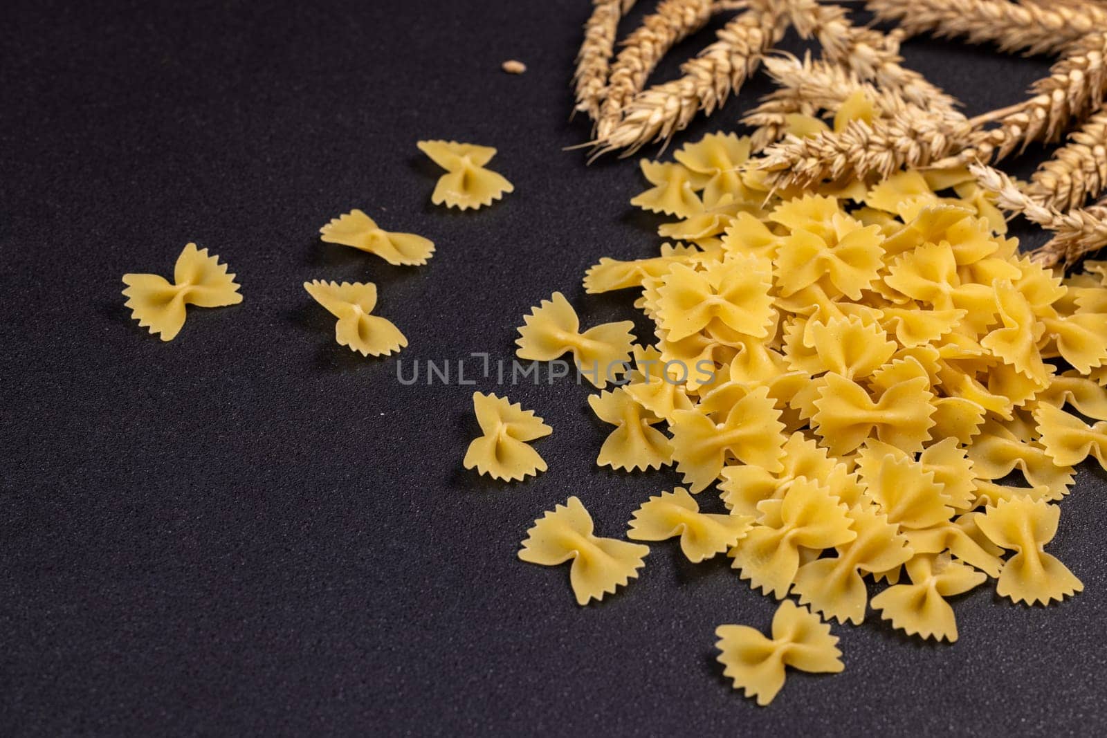 Dry farfalle pasta spilling onto a dark surface, accompanied by wheat ears, evokes a sense of rustic culinary preparation