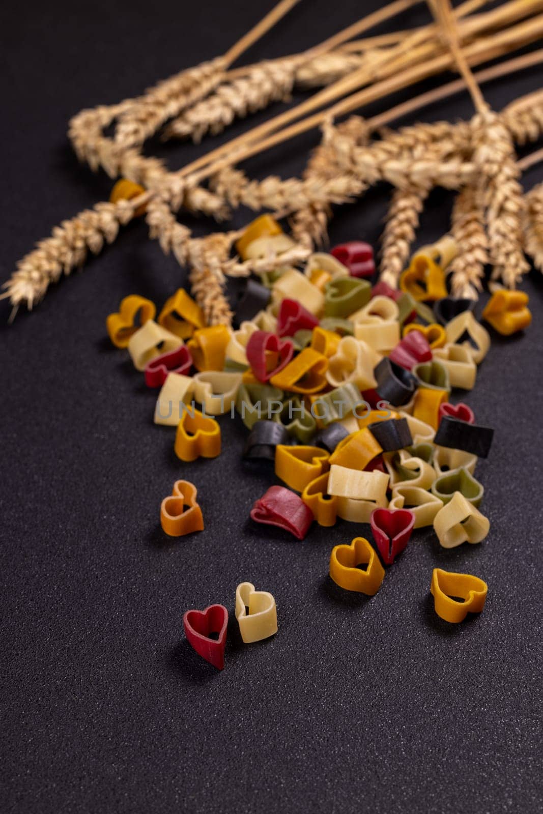 Small colorful heart shaped pasta spilling from a pile near wheat stalks on a textured black background, creating a romantic and culinary scene